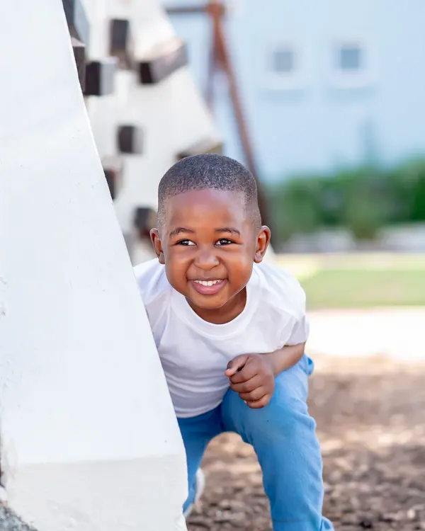 A beaming Preschool boy crouching beside a white wall on the playground