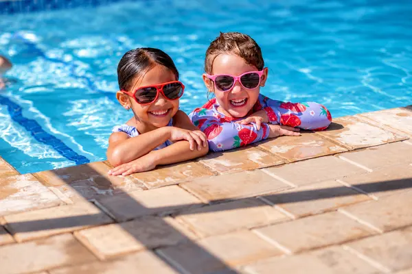 Two little girls in sunglasses grinning at the edge of the swimming pool